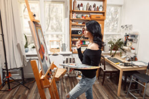 Art, creativity, hobby, job and creative occupation concept. Rear view of busy female artist sitting on chair in front of easel, painting with fingers, using white and blue oil or acrylic paint