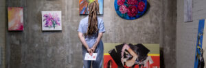 Back view of young woman standing in front of wall with artwork collection and looking at one of several abstract paintings