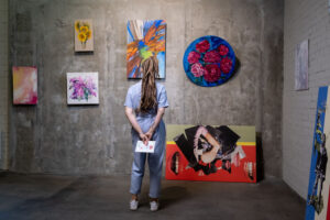 Back view of young woman standing in front of wall with artwork collection and looking at one of several abstract paintings